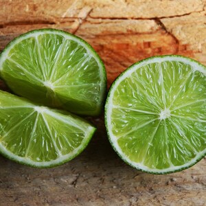 Puede incluir: Dos mitades de lima y una rodaja de lima sobre una superficie de madera. La lima es verde y tiene un interior jugoso y carnoso.