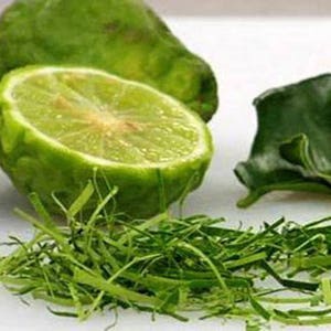 May include: Close-up of a green kaffir lime, cut in half, with green leaves and green herbs on a white surface.