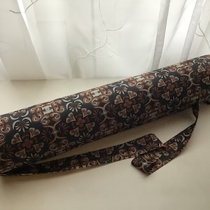 Puede incluir: Un portador de esterilla de yoga marrón y negro con un diseño estampado. El portador tiene una correa larga para llevar sobre el hombro.