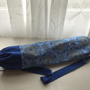Puede incluir: Una bolsa de yoga azul con un estampado de mandala colorido y un cierre de cordón azul. La bolsa tiene una correa azul larga para llevar.