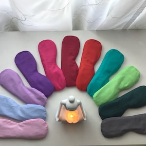 Puede incluir: Un conjunto de diez almohadas para los ojos en varios colores, dispuestas en semicírculo alrededor de un portavelas de cerámica blanca con una vela encendida en su interior. Las almohadas para los ojos están hechas de tela suave y están rellenas de un material natural.