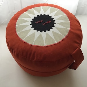 Puede incluir: Un cojín de meditación redondo de color naranja con un diseño de estrella en blanco y negro y el texto "Yoga Lover" en el centro.