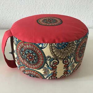 Puede incluir: Cojín de meditación redondo rojo con una tela estampada en crema y marrón con diseños de mandalas. El cojín tiene una parte superior de tela roja y un asa.