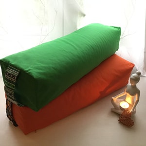 Könnte beinhalten: Zwei rechteckige Yogakissen, eines grün und eines orange, liegen übereinander. Eine kleine weiße Figur eines Menschen sitzt vor den Kissen und hält eine kleine Kerze.