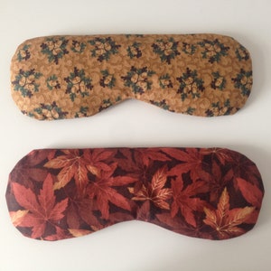 Puede incluir: Dos antifaces de tela. El antifaz superior tiene un fondo marrón con un patrón de hojas verdes y bellotas marrones. El antifaz inferior tiene un fondo marrón oscuro con un patrón de hojas de arce rojas y naranjas.