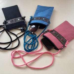 May include: Three different phone pouches with adjustable straps. The pouches are made of fabric and have embroidered designs. One pouch is black, one is blue, and one is pink.