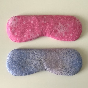May include: Two sleep masks with floral patterns. One is pink with white floral details, the other is blue with white floral details. The masks are oval-shaped and made of fabric.