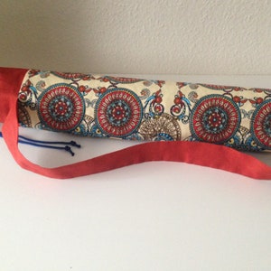 Puede incluir: Un portador de esterilla de yoga rojo y blanco con un patrón de paisley. El portador tiene un cierre de cordón rojo y una correa roja larga.