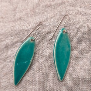 May include: A pair of teal blue ceramic earrings with silver hooks. The earrings are shaped like a leaf and have a subtle textured finish.