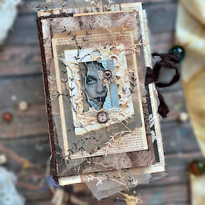 Pode incluir: Um diário artístico de técnica mista com capa castanha, apresentando papel vintage em camadas, páginas de livro rasgadas, rede preta e um retrato central. A capa inclui um adorno de borboleta e um botão circular com a letra I.