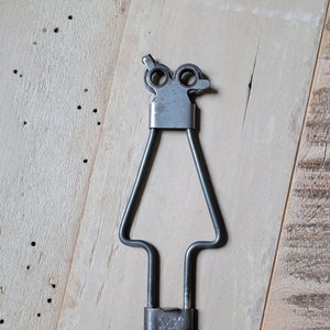 May include: A silver metal tool with a hook on the bottom and two circular handles on the top. The tool is labeled "Dritz" and "Medal" with "Made in USA" printed below.