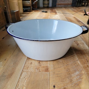 May include: White enamelware basin with a blue rim and two black handles. The basin is sitting on a wooden floor.