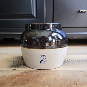 May include: A stoneware crock featuring a two-tone design with a dark brown glazed upper section and a light beige lower section. The front of the vessel displays a prominent blue number 2. The container has a wide, short neck and a rounded body.