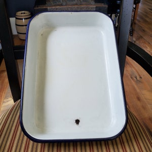 May include: Rectangular white enamelware baking dish with a dark blue rim. The interior is white, and the edges are slightly rounded. The dish has a small hole near the bottom. The dish is sitting on a striped chair.