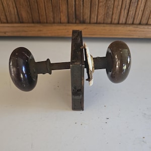 May include: An antique door knob set with dark brown, oval-shaped knobs. The set includes a rectangular metal plate and a connecting rod, showing signs of age and wear. The hardware is likely made of metal and ceramic.