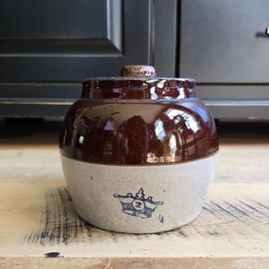 May include: A two-toned stoneware crock with a dark brown glazed upper section and a light beige lower section. The base features a blue stamped crown logo with the number 2 inside. It includes a matching lid with a small knob handle on top.