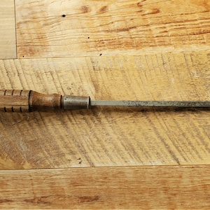 May include: A vintage screwdriver with a wooden handle and a metal shaft. The screwdriver is lying on a wooden surface.