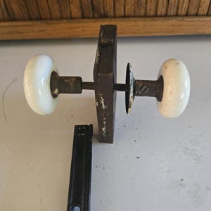 May include: Antique door hardware set featuring a rusty metal mortise lock, two white porcelain doorknobs, and a black strike plate. The hardware shows signs of age and wear.