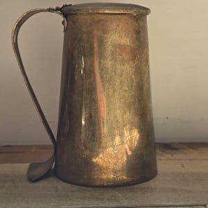 May include: A vintage brass pitcher with a handle and a worn, aged finish. The pitcher has a rounded body and a slightly flared lip.