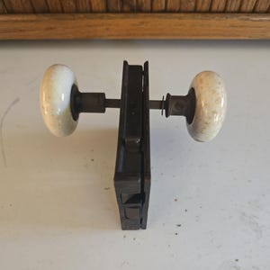 May include: Antique door hardware set featuring a black metal mortise lock with two white and tan ceramic doorknobs. The doorknobs are oval-shaped and attached to the lock with metal rods. The hardware is likely from an older home.