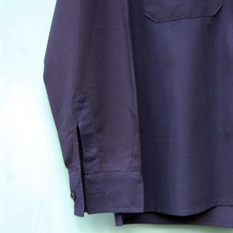 May include: A dark blue long-sleeved shirt with a single pocket on the chest. The shirt has a button-down collar and a single button cuff on each sleeve.