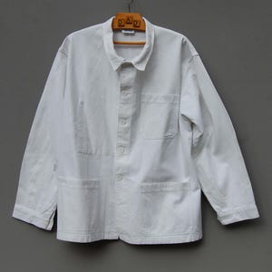 May include: A white work jacket with a collar, button-up front, and multiple pockets. The jacket is made of a sturdy fabric and hangs on a wooden hanger. The word "DAD" is visible on the hanger.