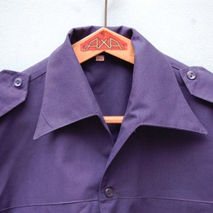 May include: A dark purple collared shirt with a button closure and a single button on each shoulder. The shirt is hanging on a wooden hanger with a label that reads "AXA" and "39".