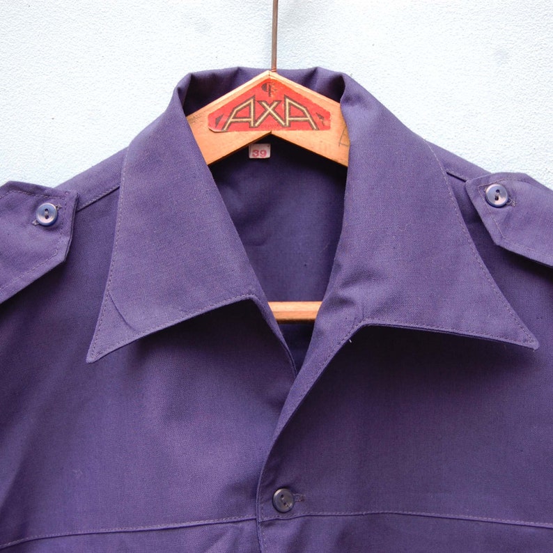 May include: A dark purple collared shirt with a button closure and a single button on each shoulder. The shirt is hanging on a wooden hanger with a label that reads "AXA" and "39".