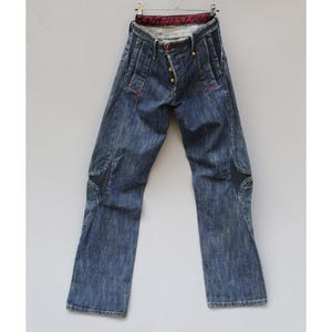 May include: Blue denim jeans featuring a button-fly closure, dark navy blue side panel inserts along the thighs and knees, and a deep red patterned fabric lining visible at the interior waistband. The jeans have a straight-leg cut and stitching.