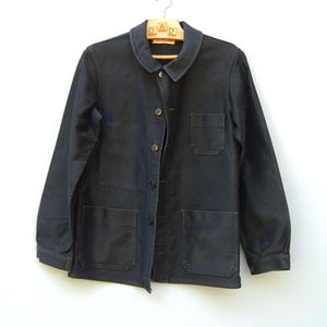 Könnte beinhalten: Marineblaue Arbeitsjacke mit klassischem Kragen, Knopfleiste und drei Vordertaschen. Die Jacke hängt an einem Holzkleiderbügel mit dem Wort "DAD" darauf. Die Jacke ist aus strapazierfähigem Stoff gefertigt.