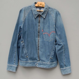 May include: A light blue denim jacket with a zip-up front closure, a collared neckline, and two front pockets. The jacket features a red stitched pocket design and a small red logo. The jacket is hanging on a wooden hanger.