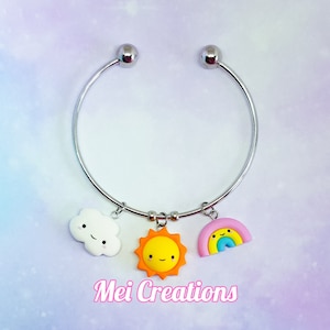 May include: A silver bangle bracelet with three charms: a white cloud with a smiley face, a yellow sun with a smiley face, and a rainbow with a smiley face.