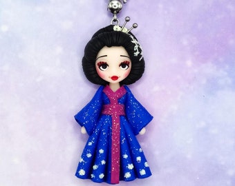 Op de afbeelding: Een blauwe en roze pop met zwart haar en een witte bloemenkroon, die een blauwe en roze kimono met witte bloemen draagt. De pop is een kettinghanger.