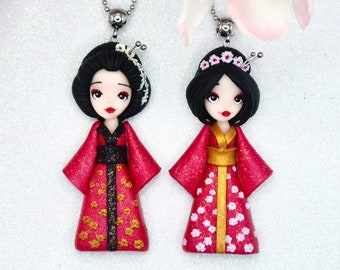 Op de afbeelding: Twee rode en gouden geisha-poppenkettingen met zwart haar en witte bloemmotieven. De poppen dragen traditionele Japanse kleding.