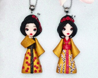 Op de afbeelding: Twee gouden kettingen met zwart haar poppen die traditionele Japanse kleding dragen. De poppen hebben rode en witte bloemenpatronen op hun jurken. Eén pop heeft een rode sjerp en de andere een witte sjerp.