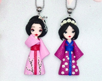 Op de afbeelding: Twee handgemaakte kettingen van polymer clay met geishapoppen die traditionele kimono's dragen. De ene pop draagt een roze kimono met witte bloemenaccenten, de andere een paarse kimono met witte stippen en een blauwe sash.
