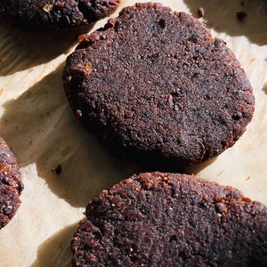 May include: Close-up of several dark brown, round cookies on parchment paper. The cookies have a rough, textured surface and appear to be freshly baked. The lighting creates shadows, highlighting the cookies' shape and texture.