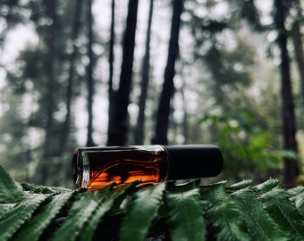 May include: A small brown glass bottle with a black cap, lying on a bed of green fern leaves. The bottle is partially filled with a dark liquid.