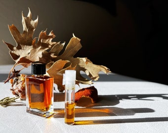 May include: Two amber glass bottles of perfume, one tall and thin, the other shorter and wider, sitting on a white surface. Dried leaves and a piece of orange peel are in the background.