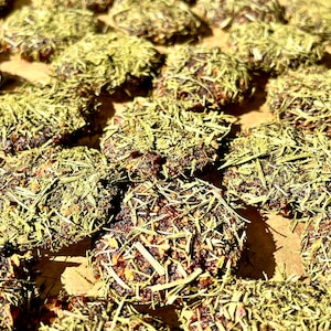 May include: Close-up of several round, dark brown food items, each generously coated with dried green herbs. The food items are arranged closely together on a light brown surface, with the herbs creating a textured, natural appearance.