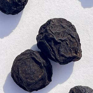 May include: Close-up of four dried black plums, also known as prunes, on a white surface.