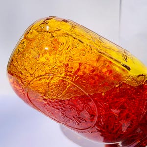 May include: A clear glass jar filled with a vibrant, layered mixture of yellow and red liquids and solids. The jar has embossed floral patterns and is tilted, showcasing the contents' color gradient. The jar is held by a hand.