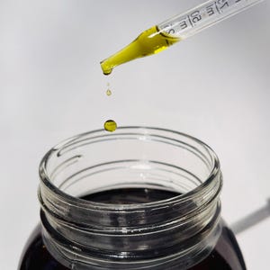May include: A clear glass jar filled with a dark liquid, with a clear glass dropper dispensing a yellow liquid. Droplets of the yellow liquid are falling into the jar. The dropper has measurement markings.