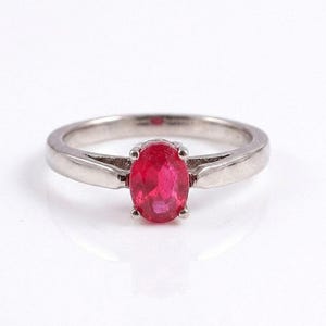 Könnte beinhalten: Ein Ring aus silberfarbenem Metall mit einem zentralen, oval geschliffenen, leuchtend roten Edelstein in einer Vier-Krappen-Fassung. Die Ringschiene ist glatt und poliert und verjüngt sich leicht zum Ansatz der Fassung des Steins.