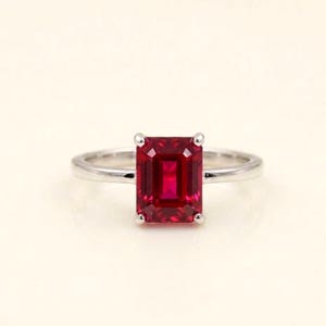 Peut inclure: Une bague argentée ornée d'une pierre précieuse rouge vif taille émeraude, fixée par quatre griffes. L'anneau est lisse et poli, présentant un design épuré qui met en valeur la forme rectangulaire et la couleur cramoisie profonde de la pierre.