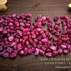 May include: A large collection of raw crimson ruby rough gemstones arranged in a cluster. A metal caliper holds a single ruby, while a jeweler's loupe and a small brass scale sit nearby. Text reads: GALAXY GEMS RAW CRIMSON RUBY ROUGH GEMSTONES | BULK LOT.