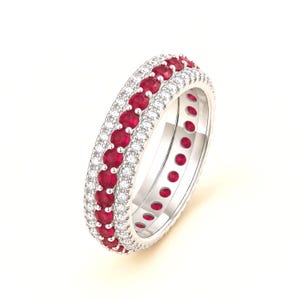 Puede incluir: Un anillo de tono plateado que presenta una fila central de rubíes rojos redondos y vibrantes, flanqueada por dos filas exteriores de pequeños diamantes blancos claros y brillantes. La banda tiene un acabado metálico pulido y diseño continuo.