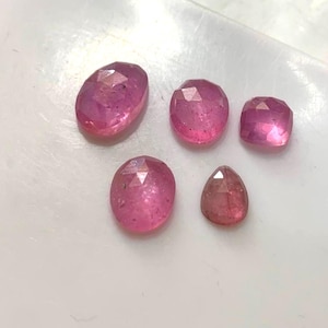 Puede incluir: Cinco piedras preciosas rosas facetadas de diversas formas y tamaños, incluyendo cortes ovalados, redondos y en forma de pera. Estas piedras translúcidas exhiben un tono magenta vibrante con inclusiones internas visibles y superficies pulidas.