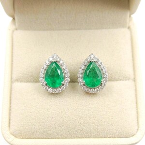 May include: A pair of pear-shaped green emerald stud earrings, each featuring a central teardrop-cut emerald surrounded by a halo of small, clear, round-cut white diamonds set in a polished silver-toned metal frame.