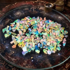 Könnte beinhalten: Eine Sammlung farbenfroher Opal-Edelsteine in einer durchsichtigen Glasschale. Die Opale zeigen eine Vielzahl von Farben, darunter Blau, Grün, Gelb und Rosa. Eine Lupe und der Text "GALAXY GEMS" sind im Hintergrund zu sehen. Die Schale steht auf einer dunklen, gemusterten Oberfläche.